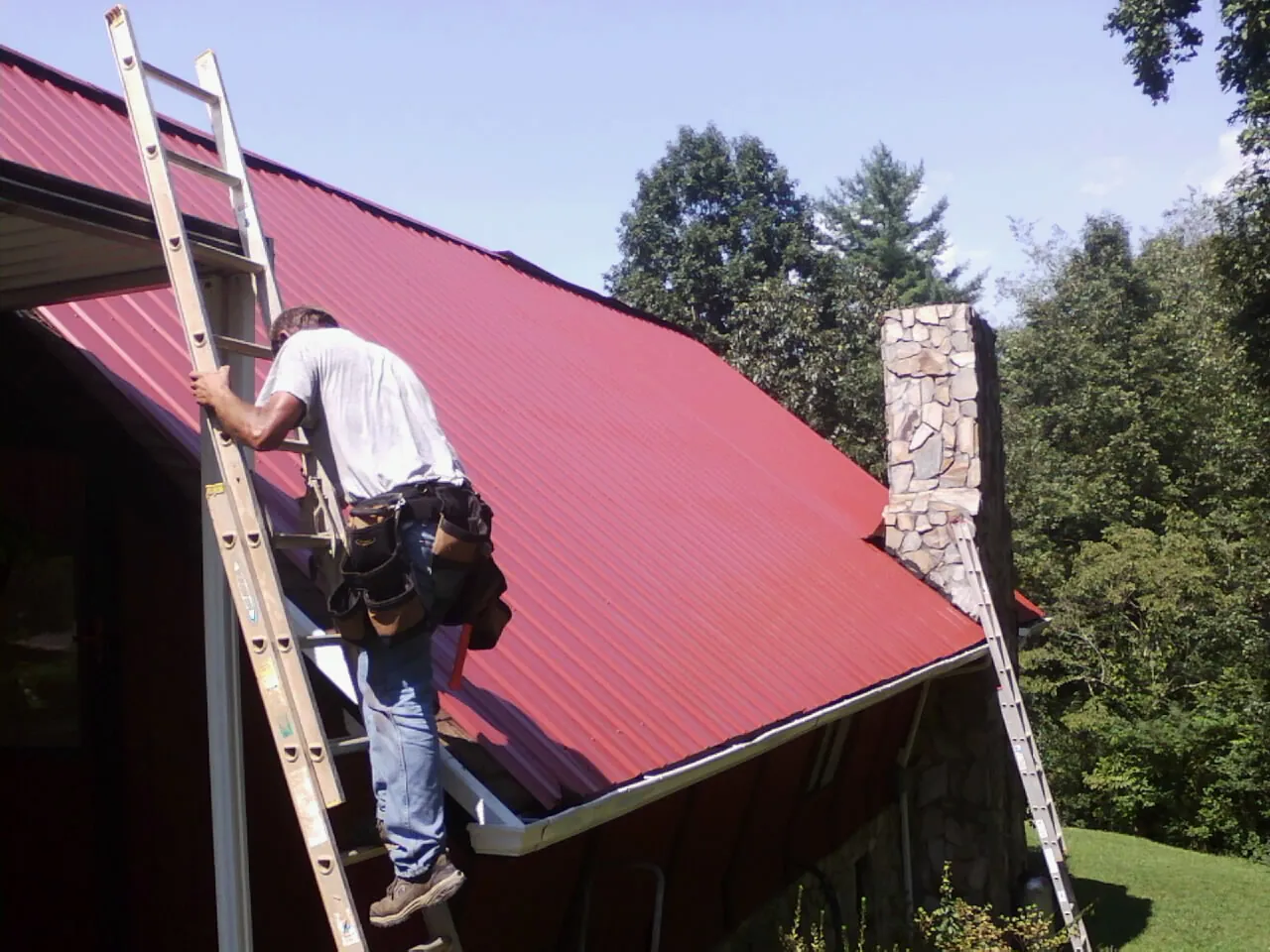 Roofer on a ladder beside a red metal roof and stone chimney — Soffit & Fascia Repair by Roofing Brothers in Owingsville